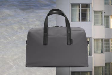 : A duffle bag in grey shown against ocean water on the left and an apartment building on the left.