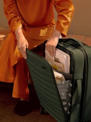 A person wearing orange clothing putting documents into the front pocket a green nylon carry-on suitcase.