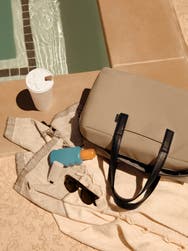 A tan bag next to a pool with a towel, coffee, sunscreen and sunglasses.