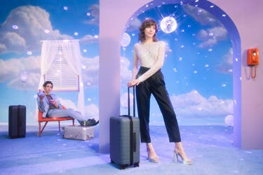 A woman carrying a coast blue colored away travel suitcase in polycarbonate. There is a man behind her, sitting on a chair with his feet resting on top of a silver aluminum suitcase and a navy polycarbonate suitcase next to him