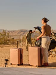 A woman standing with two checked size petal pink Away suitcases overlooking a mountain scene in Joshua tree with a man opening the trunk of a car in front of her.