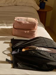 Away garment sleeve in black on a white bed with Away packing cubes in petal pink behind it.