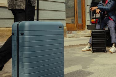 A person rolling an Away suitcase in Coast in the foreground with a second person sitting in an open trunk of a car leaning on a carry-on sized aluminum Away suitcase in black.