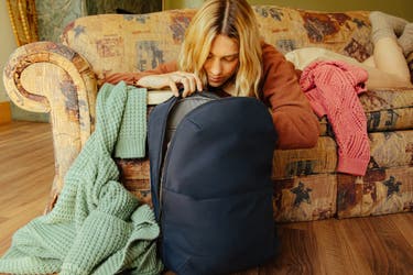 A person lying face down on a sofa while looking through an open navy Away Everywhere Zip Backpack in a cabin setting.