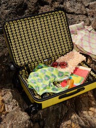 An open yellow Away Bigger Carry-On suitcase by designer Palomo Spain with various items inside on a rocky Spanish sea cliff and showing the signature inner suitcase print.