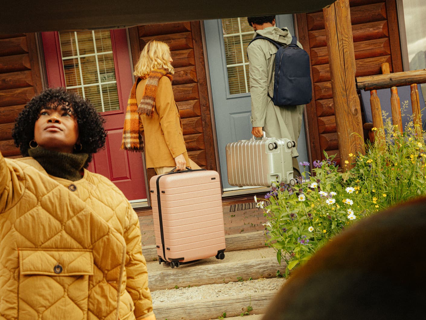 Two people in fall clothing walking up to a cabin door carrying Away suitcases and backpack, while a person in the foreground is about to close the trunk of a car.